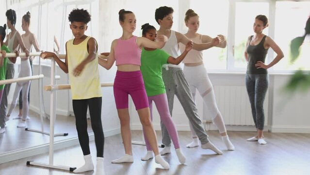 Group of concentrated teenage students of ballet school diligently practicing Demi rond de jambe par terre at barre in sunlit mirrored choreographic studio to applause of smiling female choreographer