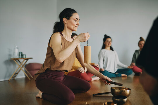 Smiling female yoga instructor beginning a meditation class using a chime and singing bowl with a group of students sitting on the floor of her studio