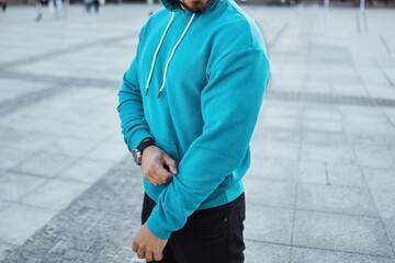 Man in a teal hoodie adjusting his sleeve, standing on an urban street