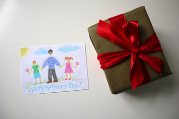 Father's Day Photo of a postcard drawn by a child. A gift of green color with a red bow. White background. The postcard shows a father walking with his daughter and son. Happy Father's Day lettering.