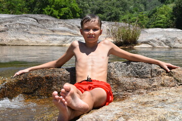 A teenage boy in orange shorts sits wet in the river. © INNA