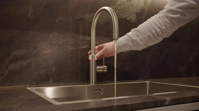 Tracking Shot Of Hand Activating point of use hot water faucet.  Hot Water flows out immediately followed by an abundance of steam.  The sink and faucet finish has a brushed stainless steel appearance
