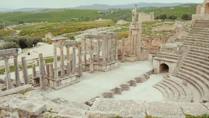 Ruins of Roman theatre in ancient settlement of Dougga in Tunisia with semi-circular orchestra, rows of stone seating and stage columns surrounded by vibrant spring greenery under cloudy sky. High