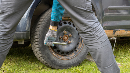 A person wearing rubber gloves is using a lug wrench to change a car tire on grass, with the vehicle's jack in place, indicating a roadside tire change or maintenance activity