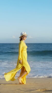 Vertical Screen: A woman in a yellow dress and hat walks on a sandy beach with waves in the background in a vertical screen format. It conveys feelings of summer, style, calmness, and peace