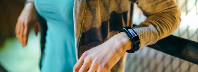 Banner of unrecognizable female wearing digital wrist band on her hand with heart rate monitor on the screen. Woman runner resting over banister with a smart sport watch checking pulse at night.