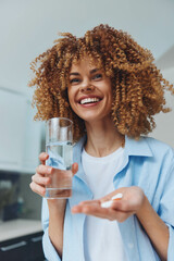 Woman with curly hair holding glass of water and pill in hand, taking medication and staying hydrated for health