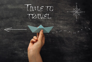 A hand holding a paper boat on a chalkboard with the words Time to Travel