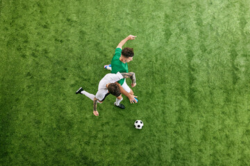 Aerial view. Dynamic photo of intense battle between two young men, soccer players for ball on green lush playground. Concept of professional sport, championship, final match, movement. Ad
