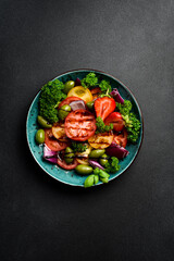 Top view of a bowl of salad made of different types of tomatoes, onions and olives, garnished with parsley and basil. On a black cement table.