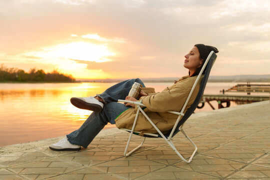 Relaxed woman sitting on deck chair near lake at sunset