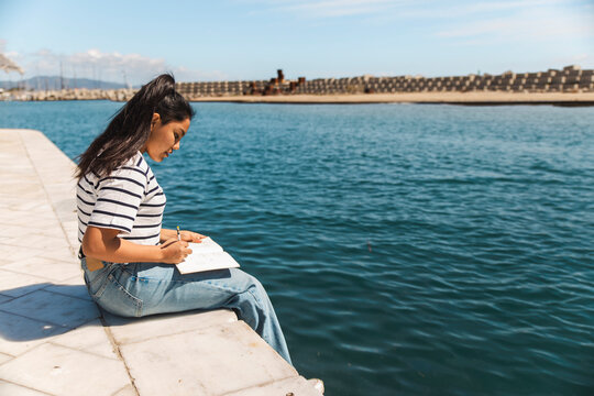 Young woman writing in book and sitting on pier by sea