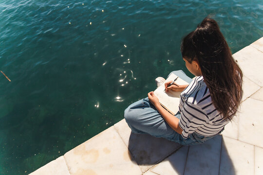 Woman writing in book by sea on sunny day