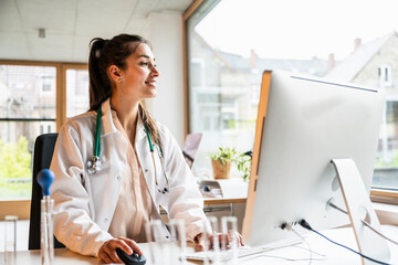 Smiling female doctor working on computer and looking through window at clinic