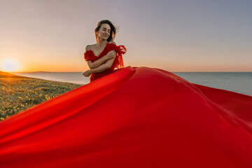 woman red dress standing grassy hillside. The sun is setting in the background, casting a warm glow...