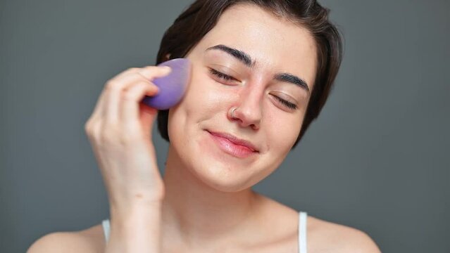 Portrait of a positive young woman doing makeup, brunette moisturizing skin with a sponge looking at the camera