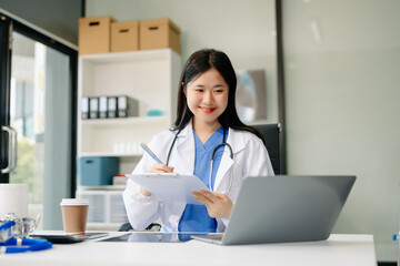 Confident young Asian female doctor in white medical uniform sit at desk working on computer....