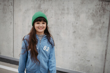 Outdoor portrait of cute young girl with beanie hat. Girl with long hair standing on playground in the city.