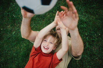 Dad lying on grass with son, laughing and having fun. Father and boy playing football, catching...