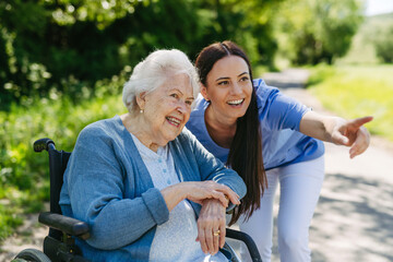 Female caregiver pushing senior woman in wheelchair. Nurse and elderly woman enjoying a warm day in...