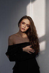 Naklejka premium Young woman with long brown hair is standing by window, wearing black off-shoulder dress. Natural makeup and long wavy hair, elegant look, natural light streaming through window