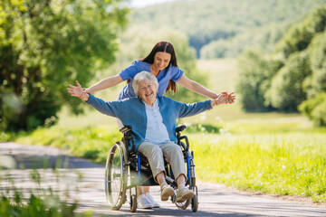 Female caregiver pushing senior woman in wheelchair. Nurse and elderly woman enjoying a warm day in nursing home, public park.