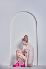 Woman in a white fur coat and pink platform heels sits by an archway in front of a white wall, posing for the camera