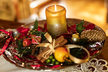 Świąteczna świeca z cynamonem, szyszkami, jabłkiem i innymi ozdobami na tle świątecznej świecącej choinki. Świąteczny nastrój © Anna