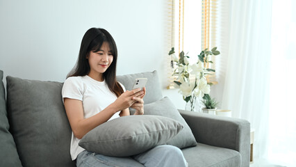 Obraz premium Charming young woman typing text message on mobile phone and relaxing on couch at home