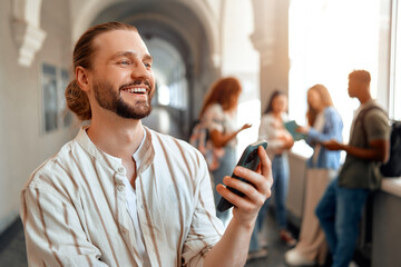 A young man wearing bright, casual clothing smiles while using a smartphone in a university hallway