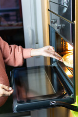 Close-up shot of woman putting a cake into the hot electric oven. in apartment. Pink sweater