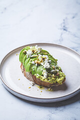 Avocado toast with feta and pistachio on plate, white marble background.