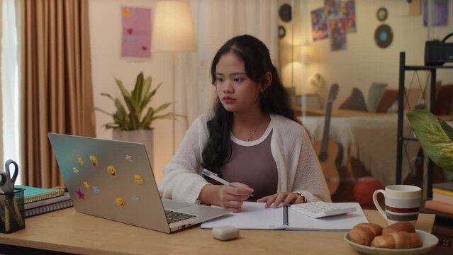 Medium shot of Asian teenage girl giving answer during online seminar when sitting in front of laptop at desk, studying remotely