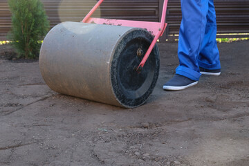 male worker in a uniform prepares a plot of land for sowing a lawn using a garden roller, leveling...