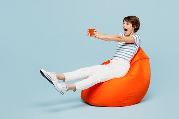 Full body young gambling woman in striped t-shirt casual clothes sit in bag chair use play racing app on mobile cell phone hold gadget smartphone for pc video games isolated on plain blue background