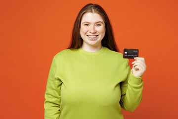 Young fat ginger chubby plus size woman wear green sweatshirt casual clothes hold in hand mock up of credit bank card isolated on red orange background studio portrait Lifestyle body positive concept