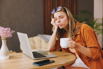 Side view young sad IT woman in orange shirt hold use laptop pc computer drink coffee sit alone at table in coffee shop cafe restaurant indoors work or study Freelance mobile office business concept