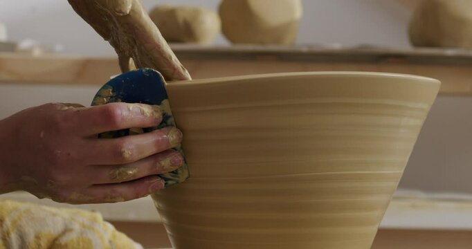 Working with a hand scraper on a big brown clay vase, pulling the scraper from the bottom to the top to get rid of material. Handheld close up shot in a daylight studio.