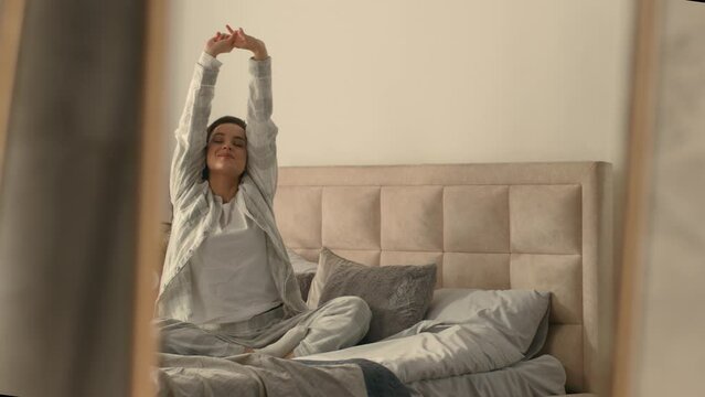 Caucasian European woman sitting on bed stretching good morning cheerful bedroom inside mirror pajamas cozy comfortable wellness carefree weekend relax resting awaken domestic holiday pleasure delight