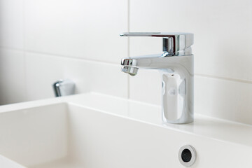 Fototapeta premium A drop of pure water dripping from the tap in the white modern bathroom. Clean tap water concept. Wise use of natural resources.