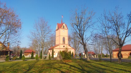 On the town street stands the historic town hall building with a clock on the tower and a metal roof with a spire. Next to it are other buildings, boulders, trees and thuja trees. Sunny autumn weather