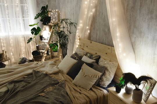Boho bed with wooden headboard, soft blankets and pillows, surrounded by light grey walls, ladder shelf with plants, and fairy lights. The room has a cozy and warm atmosphere