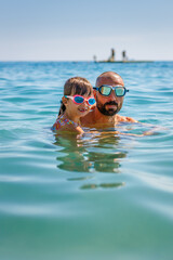 A father and his daughter, waist-deep in the calm water, both with goggles on, swimming in the sea