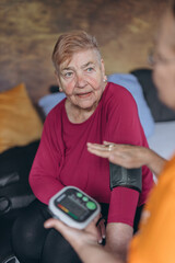 An elderly woman sits on a bed and has her blood pressure taken by a female volunteer