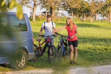 Young man is unloading electric mountain bikes, for himself and his girlfriend, lifting them from the hitch rack on the vehicle.