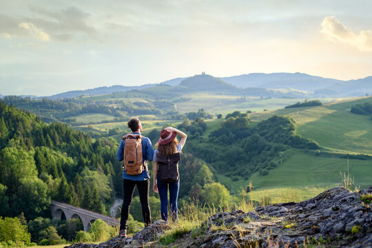 Travellers couple hiking on easy trail in nature with backpacks. Young tourist resting, enjoying breathtaking view. Summer vacation oudoors. Rear view.