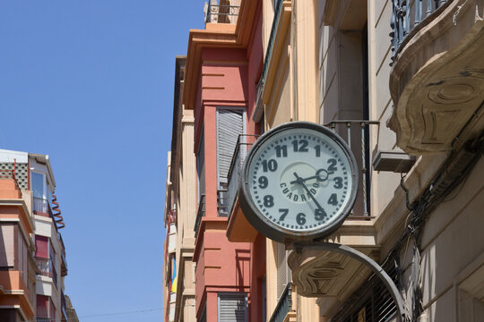 Closeup of an old analog clock that shows 2pm and is hanging from one of the buildings on a street
