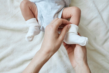 Close up of caring unrecognizable mother putting tiny socks on baby's feet baby wearing bodysuit lying on bed