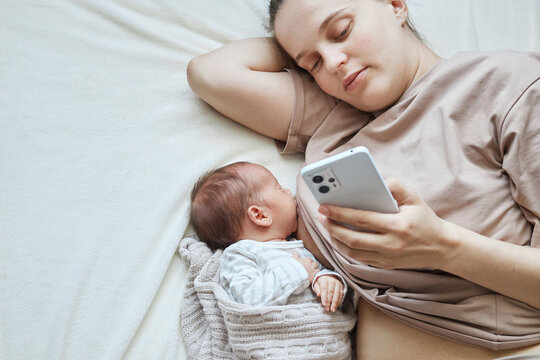 Cute infant is suckling happily on his mother's chest while mommy lying and using her mobile phone chatting with friends scrolling online - Powered by Adobe