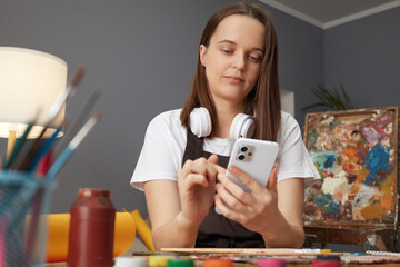 Leisure painting and craft. Concentrated Caucasian brown haired woman artist in apron working in creative workshop having break using mobile phone for online scrolling